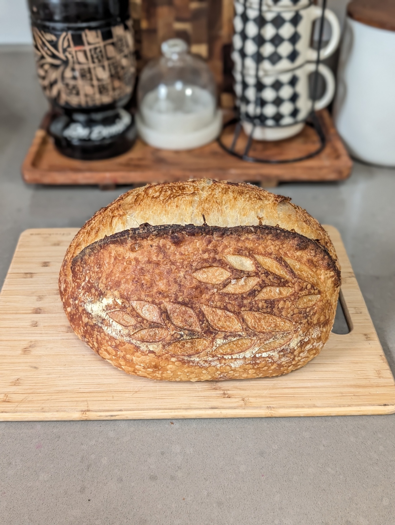 A scored oval sourdough loaf on a bamboo board, with a glass cloche and mugs behind